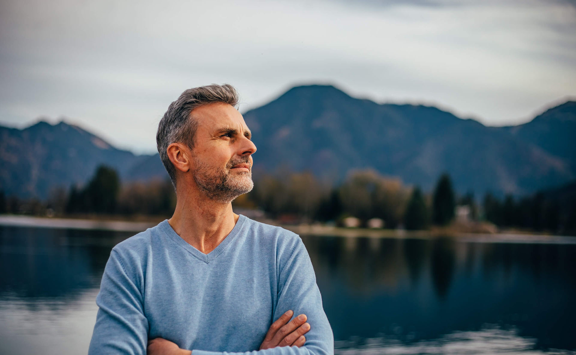 Ein mann verschänkt nachdenklich die Arme und steht vor dem Tegernsee mit Bergpanorama im Hintergrund