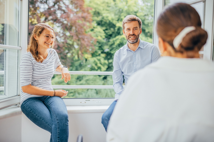 Eine Frauen und ein Mann unterhalten sich vor einem Feinster mit einer Therapeutin über Essstörungen und deren Behandlung.