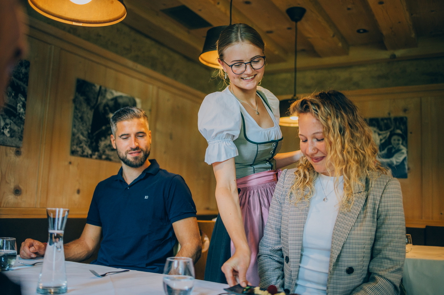 Servicekraft in Tracht serviert Gästen Getränke und Speisen an einem Restauranttisch.