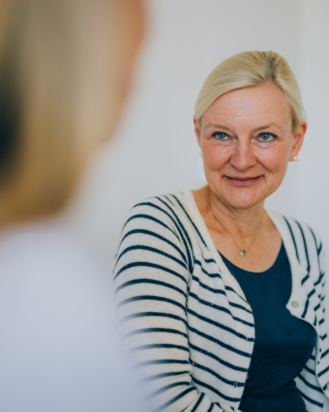 Eine blonde Frau ist sitzend im Gespräch mit einer weiteren Frau, dabei lächelt sie.