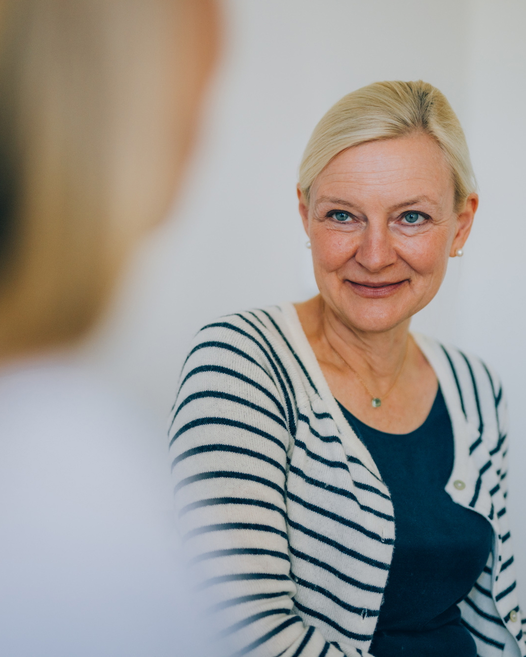 Eine blonde Frau ist sitzend im Gespräch mit einer weiteren Frau, dabei lächelt sie.