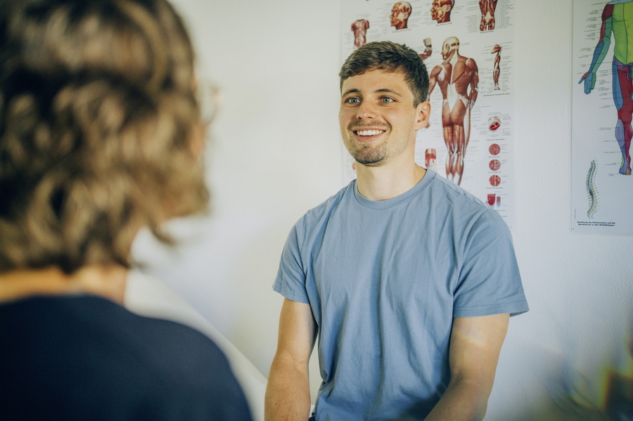 Patient sitzt auf einer Behandlungsliege in einem Behandlungszimmer und lächelt die Behandlerin an.