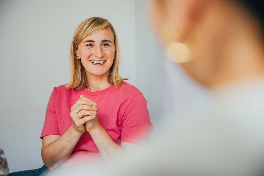 Eine Frau sitzt entspannt in einem Stuhl, ihre Hände sind gefaltet und sie lächelt ihre Gesprächspartnerin an.