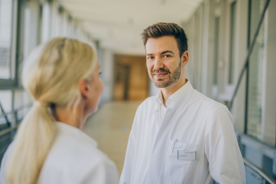 Arzt im weißen Kittel im Gespräch mit einer weiteren Ärztin auf einem Klinikflur.