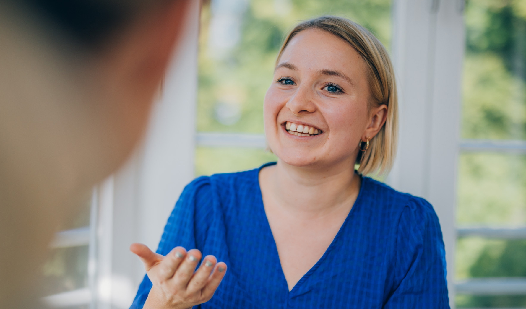 Eine Frau spricht mit einer anderen Frau in einem modernen Büro vor einem Fenster, beide wirken engagiert und freundlich. Eine gestikuliert lebhaft und lächelt.