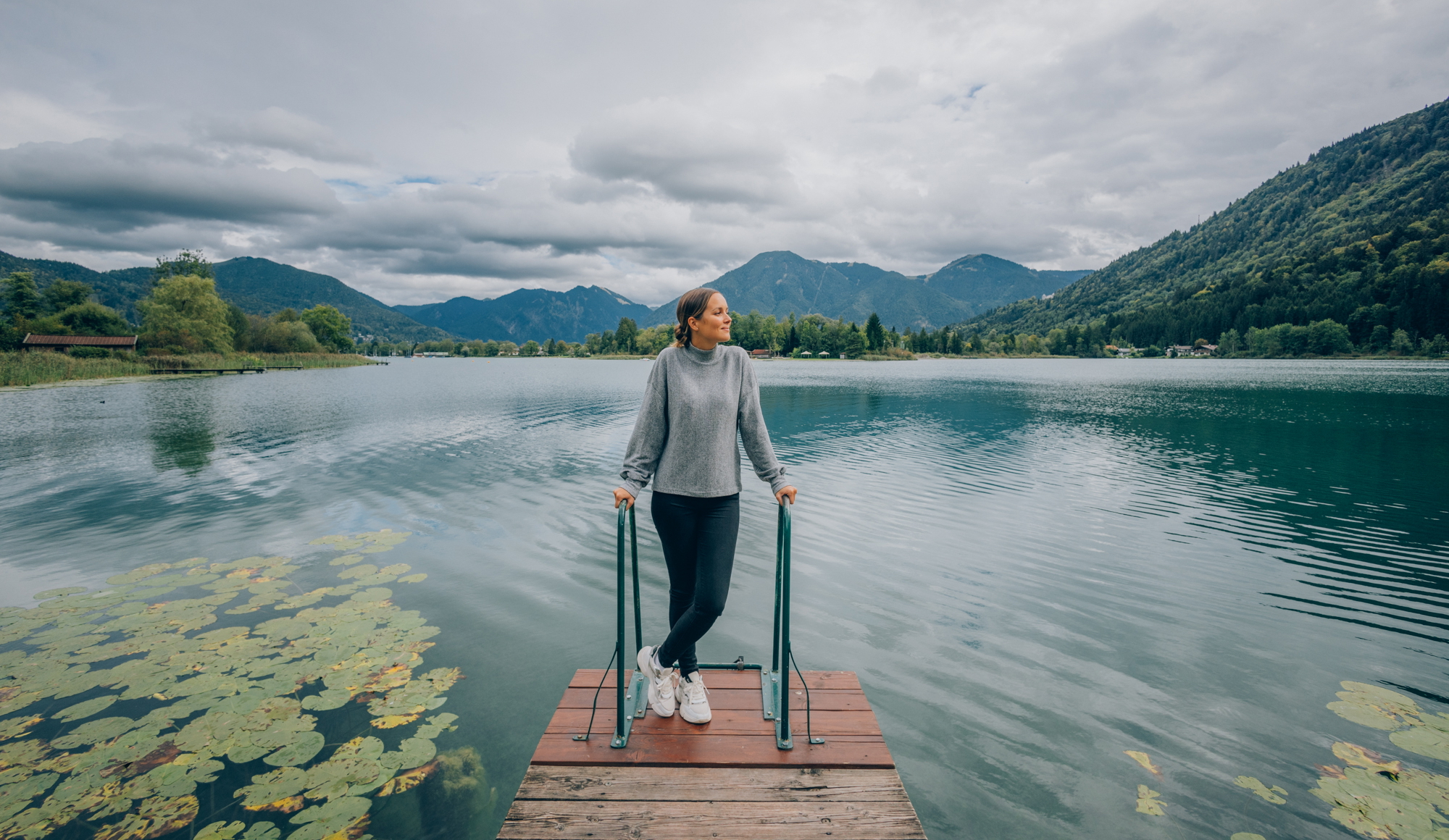 Eine Frau steht auf einem Steg am Tegernsee mit Bergpanorama im Hintergrund.
