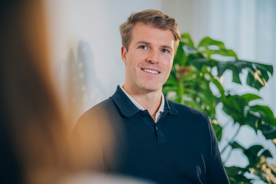 Ein lächelnder Mann sitzt in einem Büro vor einer grünen Pflanze und strahlt Freude und Zufriedenheit aus.