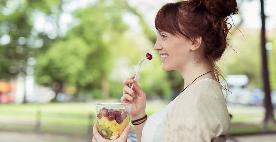 Eine Frau sitzt glücklich auf einer Parkbank und isst Obstsalat.