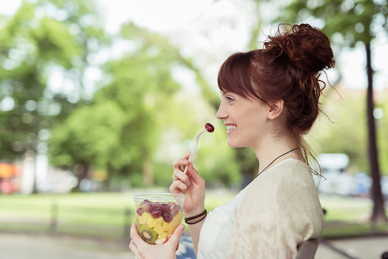 Eine Frau sitzt glücklich auf einer Parkbank und isst Obstsalat.